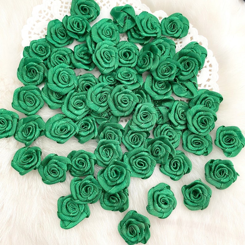 May include: A large quantity of green satin fabric roses. The roses are arranged in a random pattern on a white surface.