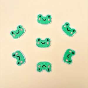 May include: A collection of seven embroidered frog patches. Each patch features a green frog with a white outline, black eyes, a smiling mouth, and pink cheeks. The patches are arranged in a circular pattern on a light yellow background.