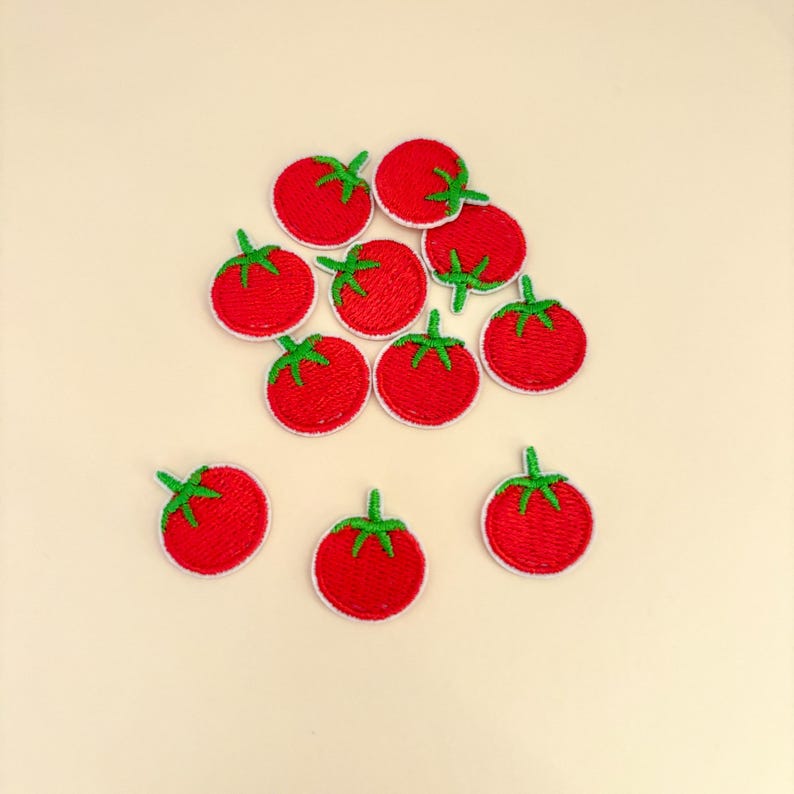 Pode incluir: Uma cole&ccedil;&atilde;o de patches de tomate vermelhos com caules verdes e bordas brancas. Os patches bordados est&atilde;o espalhados sobre uma superf&iacute;cie bege clara. Os tomates s&atilde;o de cor vermelha brilhante.