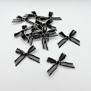 May include: A collection of small, black and gray bows with white lace trim. The bows are made of a sheer material and are arranged on a white surface. These decorative bows are suitable for crafts and embellishments.