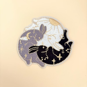 May include: Embroidered patch featuring two rabbits in a yin yang design. One rabbit is white with a daisy and the other is black with stars. Both rabbits have gold accents.