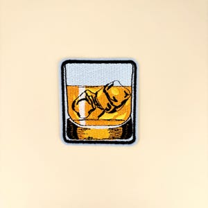 May include: Embroidered patch featuring a square-shaped design of a whiskey glass with ice cubes. The glass is filled with amber-colored liquid, and the patch has a black border with a light blue background.
