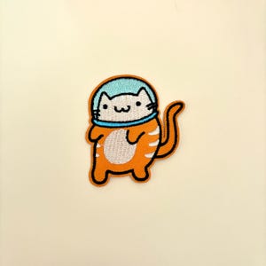May include: Embroidered iron-on patch of a white cat wearing an orange and blue astronaut suit.