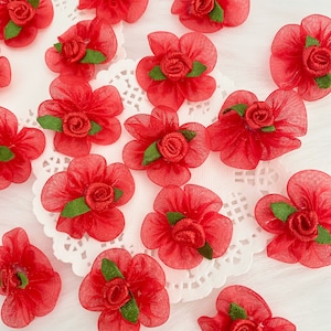 May include: A collection of red fabric flowers with green leaves. The flowers are made of tulle and have a layered, ruffled appearance. They are arranged on a white doily.