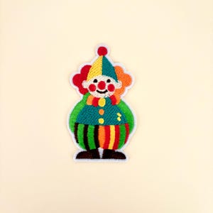 May include: A colorful embroidered patch featuring a clown with a red nose, a yellow and green pointed hat, a ruffled collar, and a green shirt with yellow buttons. The clown wears striped pants in green, orange, yellow, and black with dark brown shoes.
