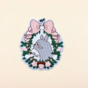 May include: An embroidered patch featuring a grey, round creature with pointed ears centered within a wreath of green leaves, pink flowers, and brown acorns. A large, light pink bow sits at the top, topped with a small white creature.