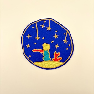May include: Embroidered patch featuring a scene from The Little Prince. The design includes a blue background with gold stars, a figure in a yellow scarf, and a red fox. The patch is circular with a gold border.