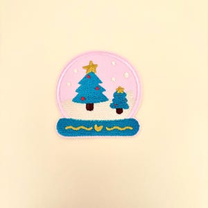 May include: Embroidered patch with a snow globe design. The globe has a pink background with a blue Christmas tree and a smaller tree, both with red ornaments. A gold star tops each tree. The base is blue with yellow accents.