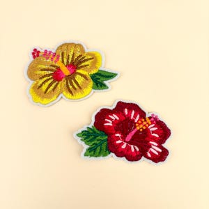 May include: Two embroidered hibiscus flower patches. One is yellow and brown with a pink center, the other is red with a pink center. Both have green leaves and a white border. The patches are on a light beige background.