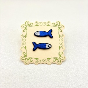 Peut inclure: Deux écussons brodés en forme de poisson avec un corps bleu et une tête blanche, présentés sur une carte carrée de couleur crème ornée d'une bordure décorative à volutes vertes. Les écussons sont disposés horizontalement l'un au-dessus de l'autre.