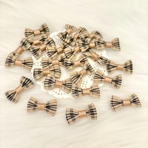 Può includere: Una collezione di piccoli fiocchi di tessuto a quadri beige e neri. Ogni fiocco ha un centro rosa pallido ed è sparso su una superficie bianca e merlettata. I fiocchi sono adatti per lavori di artigianato e abbellimenti.