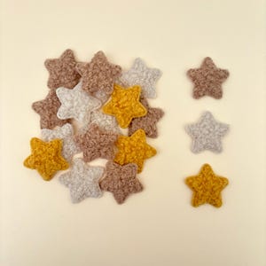 May include: A collection of star-shaped craft embellishments in various colors. The stars are in shades of gold, gray, and brown. The stars have a textured, fuzzy appearance and are scattered on a cream-colored background.