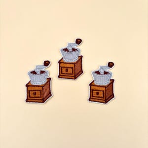 May include: Three embroidered patches of a vintage coffee grinder with a brown wooden base and a silver grinder. The patches are on a light beige background.