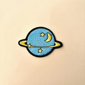 May include: Embroidered patch featuring a blue planet with yellow rings and accents. The planet has a crescent moon and three stars. The patch has a black border and is likely for clothing or accessories.