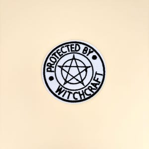 May include: A circular white patch with black embroidered text and a pentagram design. The text reads "PROTECTED BY WITCHCRAFT" around the edge of the circle, with a five-pointed star in the center.