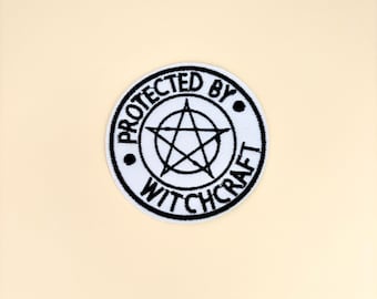 Puede incluir: Un parche circular blanco con texto bordado en negro y un diseño de pentagrama. El texto dice "PROTECTED BY WITCHCRAFT" alrededor del borde del círculo, con una estrella de cinco puntas en el centro.