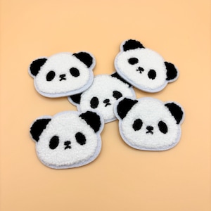 May include: Five fluffy panda face patches. Each patch features a white face with black ears and eyes, and a light blue border. The patches are arranged on a light orange background.