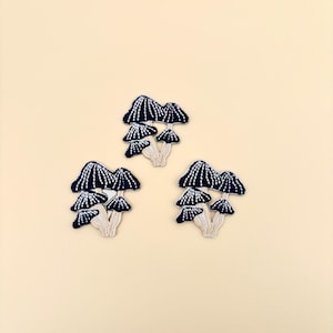 May include: Embroidered mushroom patches in navy blue and white. The mushroom caps are navy blue with white vertical lines, and the stems are beige. The patches are arranged in a group of three on a light beige background.