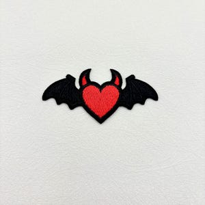 May include: Embroidered patch featuring a red heart with devil horns, framed by black bat wings. The patch is designed with a detailed stitch pattern, creating a textured appearance. The patch is likely for clothing or accessories.