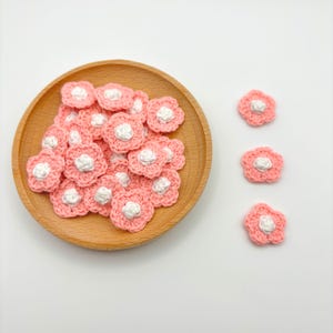 May include: A collection of pink crochet flower appliques with white centers, displayed in a wooden bowl and scattered on a white surface. These handmade floral embellishments are ideal for crafting and adding a delicate touch to various projects.