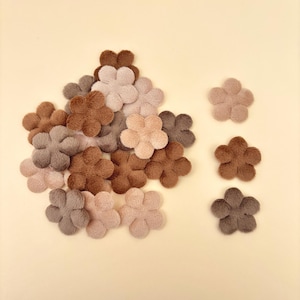 May include: A collection of small, plush, five-petal flower appliques in various shades of brown, beige, and gray. The flowers are scattered across a light yellow background, with some grouped together and others isolated.