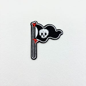May include: Embroidered pirate flag patch featuring a black flag with a white skull and crossbones design. The flag is waving on a black and white striped pole, with red heart accents. The patch is on a white background.