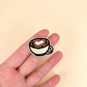 May include: Embroidered patch featuring a coffee cup with a heart-shaped design in the foam. The cup is white with a brown coffee design and a black outline. The patch is small and detailed, suitable for clothing or accessories.