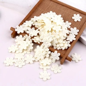 May include: A large pile of white fabric flower embellishments, scattered on a white surface. The flowers are small and delicate, with a slightly raised texture.