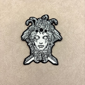 May include: Black and white embroidered patch featuring a Medusa design. The patch depicts a woman's face with snakes for hair, tears, and a rose at the base. The patch has a black border and is set against a neutral background.