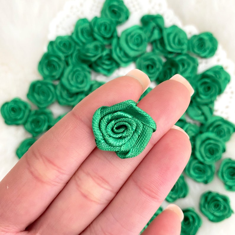 May include: A close-up of a hand holding a green satin rose. The rose is made of fabric and is about 1 inch in diameter. There are many other green satin roses in the background.