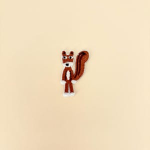 May include: Embroidered patch featuring a cartoon squirrel. The squirrel has a brown body, a fluffy brown tail, and white accents on its face, chest, and feet. The patch is designed with a playful, whimsical style, suitable for clothing or accessories.