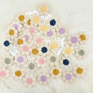 May include: A collection of crocheted flower embellishments in various colors, including pink, purple, yellow, blue, and green. The flowers are all approximately 1 inch in diameter and have a delicate, handmade appearance.