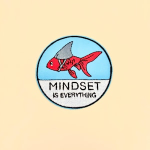 May include: Embroidered patch featuring a red goldfish wearing a shark fin, set against a light blue circle. The text "MINDSET IS EVERYTHING" is displayed below the fish in white lettering on a black background. The patch is on a cream-colored background.