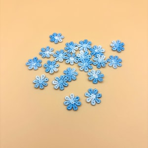 May include: A collection of small, blue and white embroidered flower appliques. The flowers are in various shades of blue, with white accents, and are scattered on a light tan background. These delicate floral embellishments are suitable for craft projects.