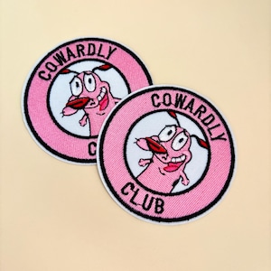 May include: Two round, pink embroidered patches featuring the cartoon dog Courage from the show *Courage the Cowardly Dog*. The patches have a white center with the dog's face and the words "COWARDLY CLUB" and "COWARDLY" in black text.