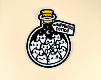 May include: Embroidered patch featuring a black and white illustration of a potion bottle filled with cat faces. The bottle has a yellow stopper and a tag that reads "HAPPINESS POTION". The patch is outlined in white.