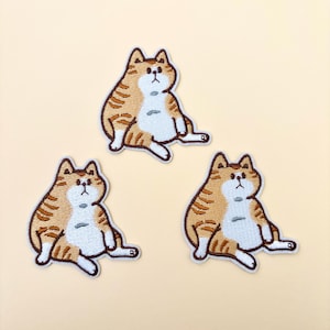 May include: Three embroidered iron-on patches of a cartoon orange tabby cat sitting with its head tilted up. The cat is outlined in white and has brown stripes.