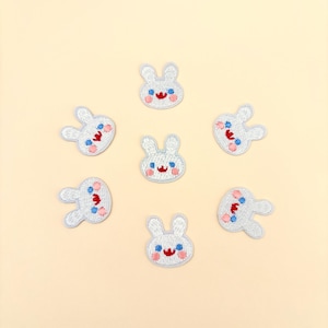 May include: Embroidered patches featuring a cartoon bunny design. Each patch is white with light blue outlining, a red mouth, and blue and pink accents. The patches are arranged in a circular pattern on a light yellow background.