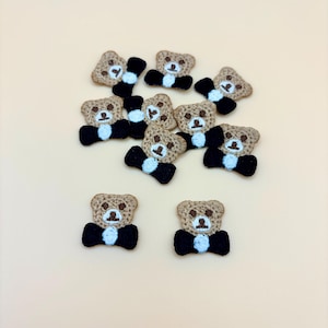 May include: A collection of small, embroidered teddy bear patches. Each bear has a tan body, a black bow tie, and a light blue shirt collar. The patches are scattered on a light beige surface.