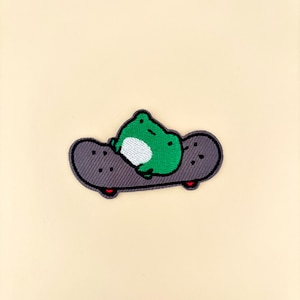 May include: Embroidered patch featuring a green frog with a white belly lying on a gray skateboard. The skateboard has black wheels with red accents. The patch is set against a cream-colored background.