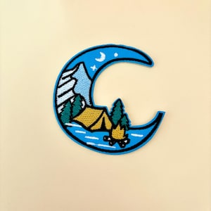 May include: An embroidered patch featuring a crescent moon with a blue sky, white stars, and a mountain range. A yellow tent is set up in a clearing with green trees and a campfire in front of it.