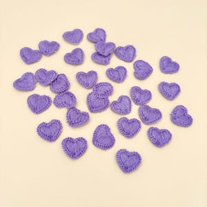 May include: A collection of small, lavender-colored, embroidered heart-shaped appliques. Each heart is crafted with a textured stitch, creating a raised surface. The appliques are scattered on a light beige background, suitable for crafts and embellishments.