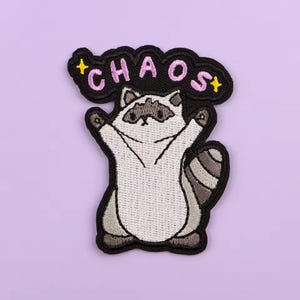 May include: Embroidered patch featuring a cartoon raccoon with its arms raised. The patch has the word "CHAOS" in pink letters above the raccoon, with yellow star accents. The patch is outlined in black and set against a lavender background.