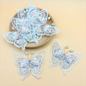 May include: Delicate white lace butterfly appliqués with pink and blue embroidered details and pearl accents. Some butterflies are arranged on a wooden plate. The butterflies have gold wire antennae.