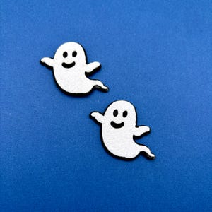 May include: Two small, white, flat ghost-shaped cutouts with black outlines and simple black facial features, including two oval eyes and a curved mouth. Each ghost has two outstretched arms and a trailing tail, presented as a pair of identical figures.