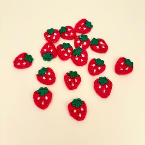 May include: A collection of small, crocheted strawberries. Each strawberry is red with white accents and a green stem. The strawberries are scattered on a light yellow background.