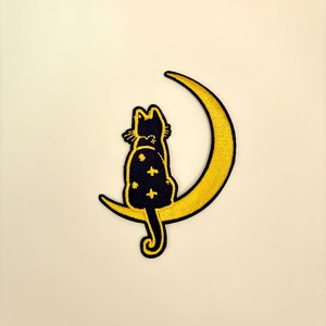 May include: A black cat with a starry pattern sits on a crescent moon, both embroidered in yellow thread.