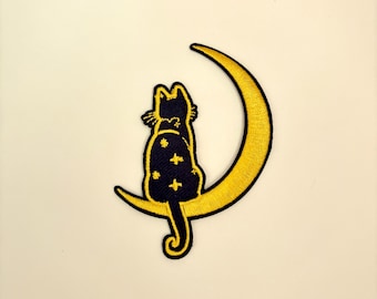 May include: A black cat with a starry pattern sits on a crescent moon, both embroidered in yellow thread.