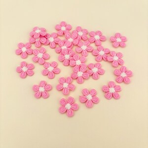 May include: A collection of pink fabric flowers with white centers. The flowers are scattered on a light beige background.