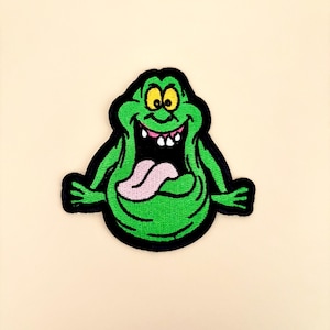 May include: A green embroidered patch featuring a cartoon ghost character with a wide open mouth and a pink tongue. The ghost has yellow eyes, a black outline, and is smiling. The patch is likely for clothing or accessories.